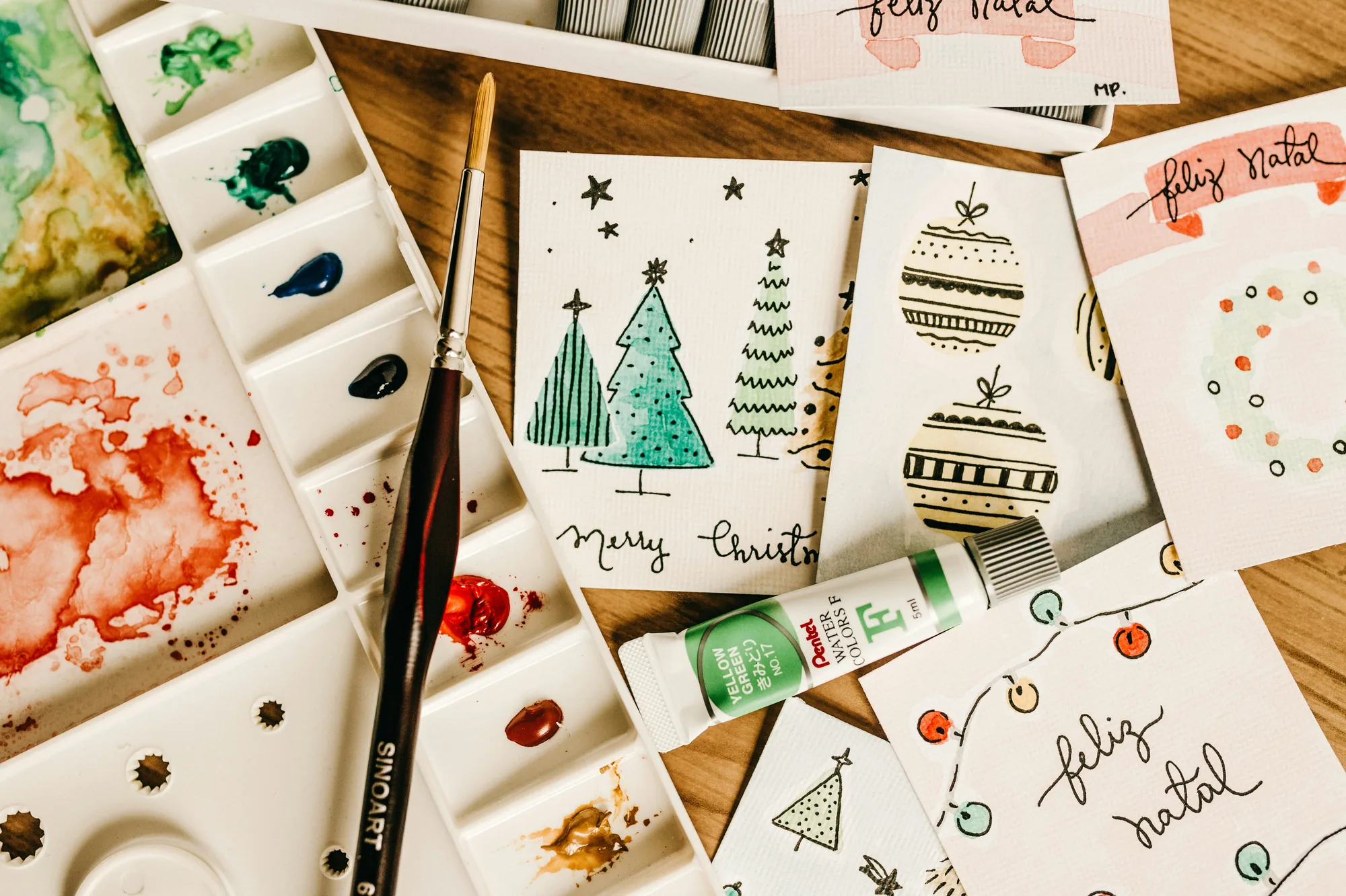 Hand-lettered watercolor holiday cards painted with trees, ornaments and string lights, beside a paint palette and brush