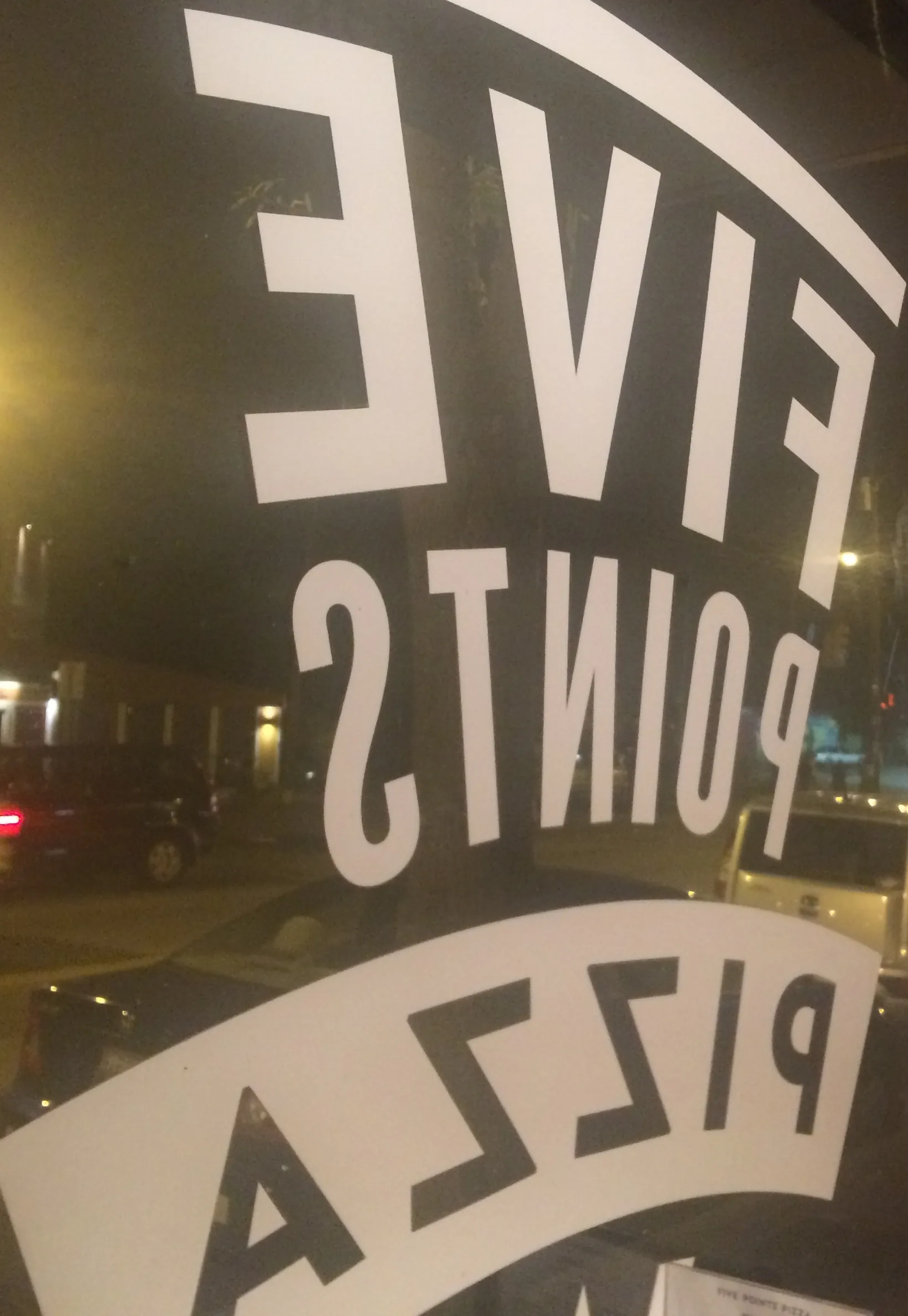 Five Points Pizza lettered on a window, seen in reverse from inside at night, with parked cars and streetlights beyond