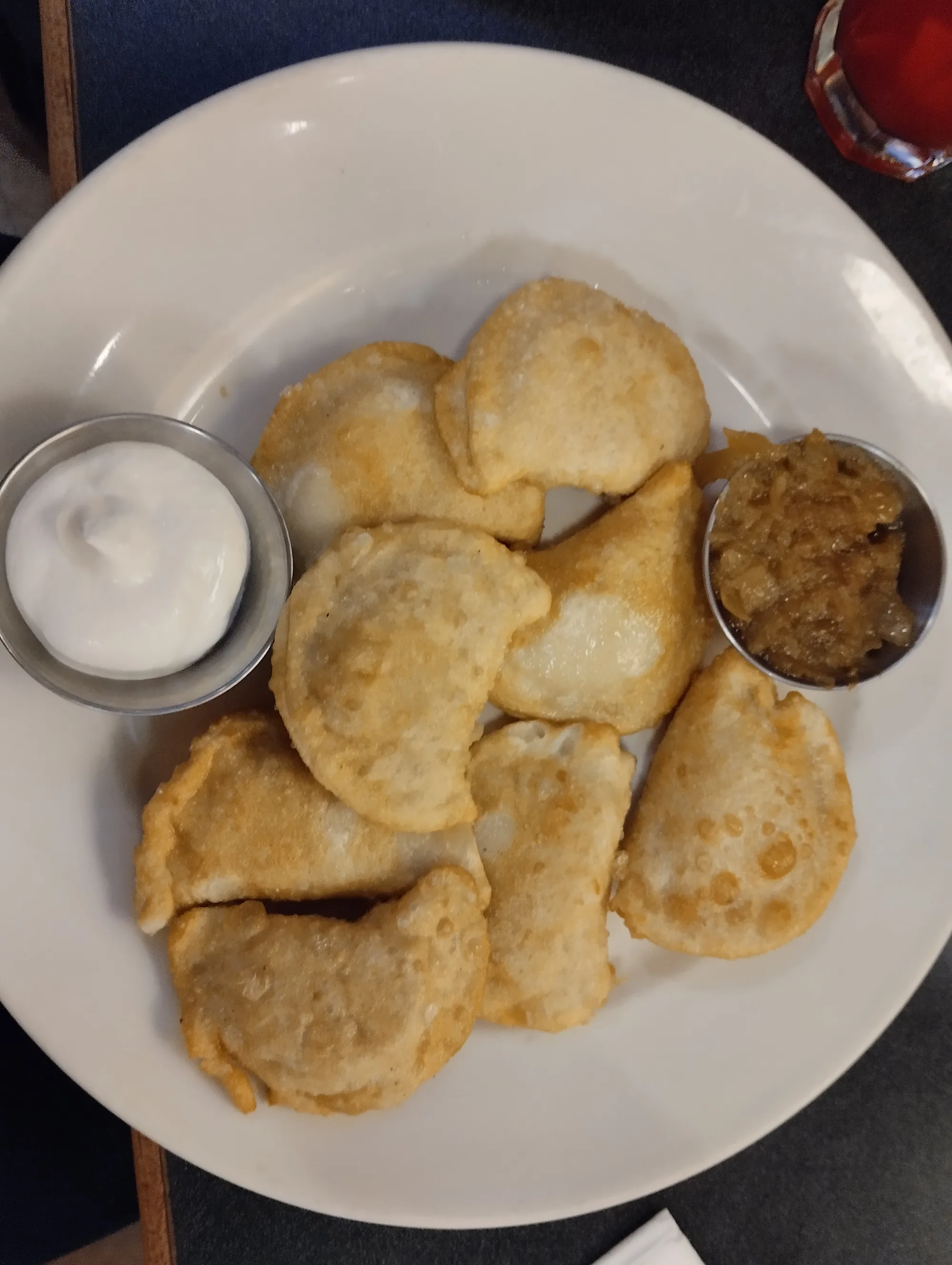 Fried pierogi on a white plate with small cups of sour cream and fried onions